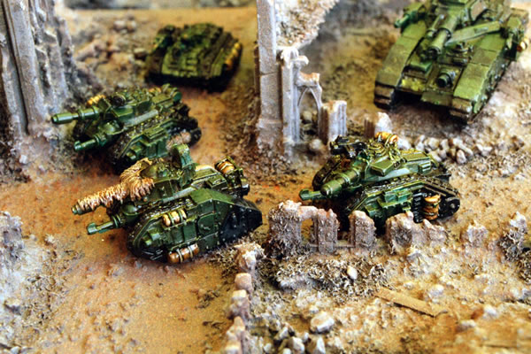 Imperial Leman Russ tanks supported by Chimeras and a Baneblade move through the ruins of an Imperial City.